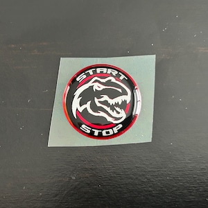 May include: A black and red round sticker with a white dinosaur head graphic and the words "START STOP" in white lettering.