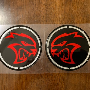 May include: Two black and red round car emblems with a white outline. Each emblem features a red silhouette of a snarling, stylized animal head.