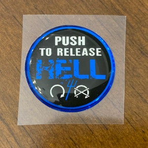 May include: A round sticker with a black background and blue border. The text "PUSH TO RELEASE HELL" is in white and blue. Below the text are two white symbols. The sticker is on a clear backing.