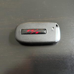 May include: A black car key fob with a rectangular red "R/T" emblem. The key fob is oval-shaped with a textured surface and a small button on the side. The background is a dark wooden surface.