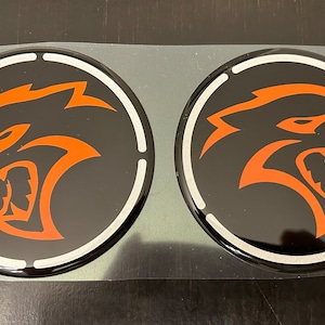 May include: Two black and orange car emblems with a circular design. Each emblem features a snarling orange cat head within a black circle, outlined with a white border. The emblems are on a gray backing.