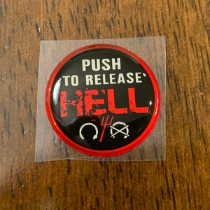 May include: A red and black round button with the text "PUSH TO RELEASE HELL" in white lettering. The button also features a black horseshoe, a black pitchfork, and a black circle with a diagonal line through it.