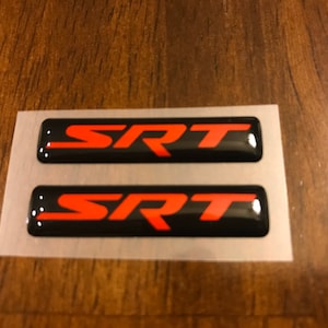May include: Two black and red SRT emblems on a white backing. The emblems are raised and have a glossy finish.