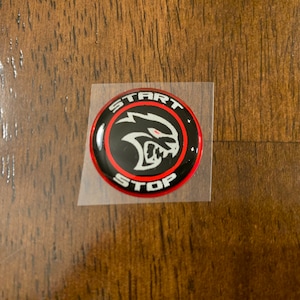 May include: A black and red round sticker with a white silhouette of a snarling animal head. The sticker says "START STOP" around the edge.