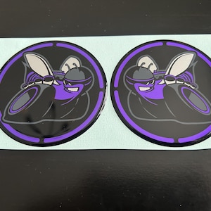 May include: Two circular black and purple decals featuring a stylized cartoon hornet with sunglasses. The hornet is in a black circle with a purple border. The decals are on a light blue surface.
