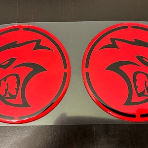 May include: Two red round stickers with a black silhouette of a snarling animal head. The animal has a pointed snout, sharp teeth, and a prominent brow.