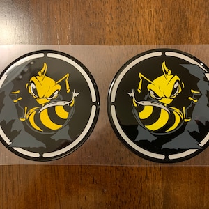 May include: Two black and white round stickers with a yellow and black bee graphic. The bee is depicted with a menacing expression and is surrounded by a black and white circle.