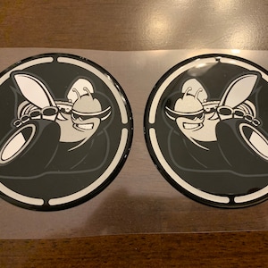 May include: Two black and silver round decals with a cartoon bee wearing sunglasses and holding a tire. The bee is in a black and white circle with a silver ring around the outside.