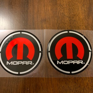 May include: Two black and silver round stickers with a red Mopar logo. The logo is a stylized letter M with a circle around it. The text "Mopar" is below the logo.