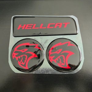 May include: A set of three black automotive emblems with red accents. The top emblem reads "HELLCAT" in red, and the two circular emblems feature a red snarling cat design.