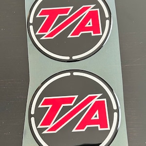 May include: Two black and white round stickers with a red "T/A" logo in the center. The stickers are on a clear plastic sheet.