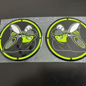 May include: Two circular stickers featuring a stylized cartoon hornet in black, gray, and neon green. The hornet is wearing sunglasses and a helmet. The stickers are on a clear backing.