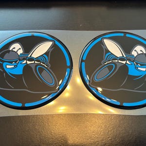 May include: Two circular black and blue decals featuring a stylized cartoon hornet with a blue helmet and wings. The hornet is set against a black background with a blue border. The decals are on a clear backing.