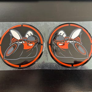May include: Two circular black and orange decals featuring a stylized cartoon hornet with a helmet and goggles. The hornet is set against a black background with an orange border. The decals are on a clear backing.