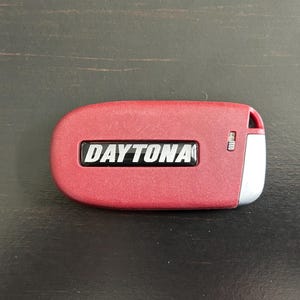 May include: A red car key fob with a silver trim and the word "DAYTONA" printed in silver letters on a black background.
