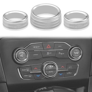 May include: Three silver car knob covers with a textured finish. The knobs are round and have a raised lip around the edge. The knobs are designed to fit over the existing knobs on a car's dashboard.