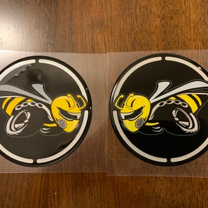 May include: Two black and yellow bee emblems with a white outline. The bee is stylized with a helmet and wings. The emblems are on a white background.
