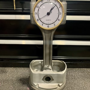 May include: A unique clock made from a silver-colored engine piston. The clock face is white with black numerals and hands, and a gold-colored bezel. The word "QUARTZ" is printed on the clock face. The clock is a creative decorative item.