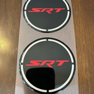 May include: Two black round car coasters with a white ring around the edge and the word "SRT" in red letters.
