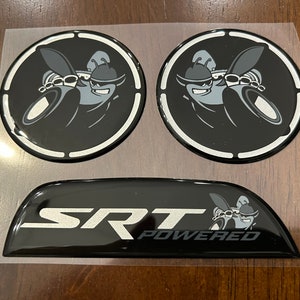 May include: Three black and silver car emblems. Two round emblems feature a cartoon character with a black and white design. The third emblem is a curved shape with the text "SRT POWERED" in silver lettering.
