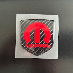 May include: A black and red Mopar emblem with a carbon fiber pattern. The emblem is a shield shape with a red M in the center.