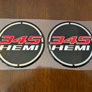 May include: Two black and silver round car emblems with a white outline. The emblems have the text "345 HEMI" in red and white lettering.