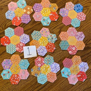 May include: Nine fabric coasters, each composed of seven hexagonal patches sewn together in a flower shape. The patches feature various floral and geometric patterns in bright colors including red, yellow, blue, pink, and orange. A small white card with the number 1 is included.