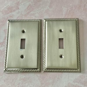 May include: Two rectangular light switch wall plates made of brushed silver-toned metal. Each plate features a central vertical toggle switch opening, two screw holes for mounting, and a decorative rope-style border along the outer edges.