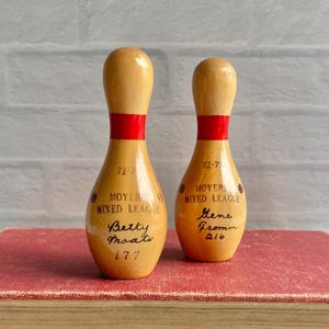 May include: Two light-colored wooden bowling pins with a red stripe around the neck. Each pin features the text 72-73, MOYERS MIXED LEAGUE, and a handwritten name and score, specifically Betty Moats 177 on one and Gene Fromm 216 on the other.