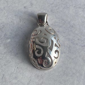 May include: A silver pendant with an oval shape and a decorative openwork design. The pendant has a small loop at the top for a chain. The design features swirling patterns. The pendant is displayed on a gray surface.