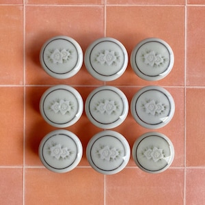 May include: Nine white ceramic cabinet knobs with a floral design and a dark gray border are arranged on a peach-colored tile surface. The knobs are round and have a raised floral pattern in the center.