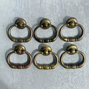 May include: Six antique brass drawer pulls with a ring handle design. The pulls have a round backplate and a curved handle. The brass has a warm, aged patina, adding to their vintage charm. Ideal for furniture restoration or adding a classic touch.