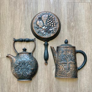 May include: Three decorative wall plaques in a copper and black finish. One plaque is a round skillet with a fruit relief, one is a rounded kettle with a floral relief, and one is a tall coffee pot with a floral relief and a side handle.