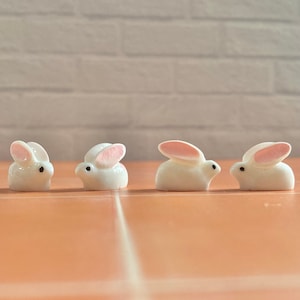 May include: Four white ceramic bunny figurines with pink ears. The bunnies are arranged in a row on a light orange surface.