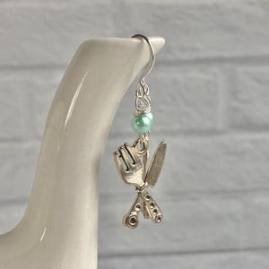May include: A single silver earring featuring a fork and knife charm, accented with a small, light green bead. The earring hangs from a silver hook. The background is a white brick wall.