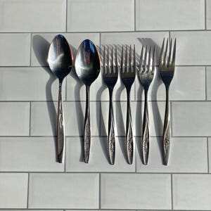 May include: A set of six stainless steel silverware pieces, including two spoons and four forks. The silverware has a floral design on the handles and is arranged on a white tiled surface.