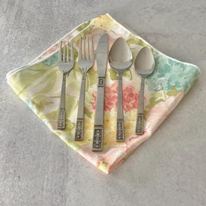 May include: A set of vintage silverware, including forks, a knife, and spoons, arranged on a floral patterned napkin. The silverware has decorative handles. The napkin features a pastel color palette of pink, yellow, and green.