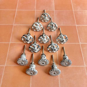 May include: A collection of decorative cabinet knobs with a distressed white and black finish. The knobs have a variety of floral and geometric designs, with metal stems. The knobs are arranged on an orange tiled surface.