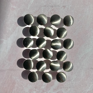May include: A collection of twenty-four silver-toned, domed buttons arranged in a grid pattern. The buttons have a metallic sheen, reflecting light and creating highlights. The background is a light pink, textured surface.