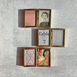 May include: A collection of small, hinged, gold-framed table card holders. Some hold vintage images of people, while others feature patterned paper in pink and white. One card displays the text "Table 7".