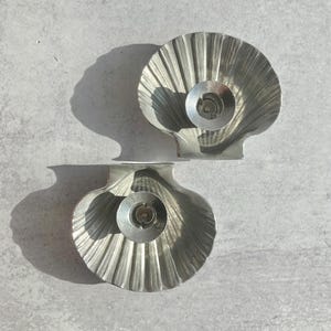 May include: Two silver-colored seashell-shaped candle holders. Each holder has a central circular indentation for a candle. The holders are made of a ridged metal and sit on a light gray surface.