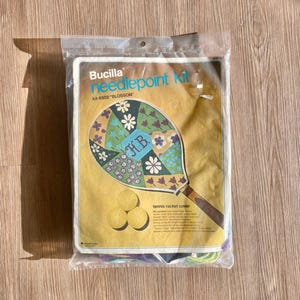 May include: A Bucilla needlepoint kit for a tennis racket cover, titled "Blossom." The kit features a colorful tennis racket design with floral and geometric patterns, three tennis balls, and the initials "H.B." in the center.
