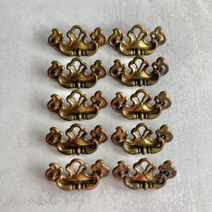 May include: A set of ten matching brass drawer pulls arranged in two vertical columns. Each pull features a symmetrical, ornate design with a central arched element, curved side flourishes, and two mounting points for attachment to furniture.