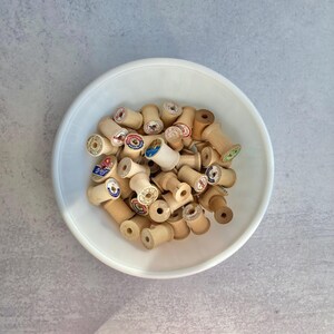 May include: A white bowl filled with a collection of wooden spools. The spools vary in color, with some displaying colorful labels. The spools are arranged in a random pattern, creating a visually appealing arrangement. The image is taken from a top-down perspective.