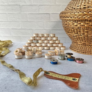 May include: A wicker basket with a lid sits next to a pile of white thread spools. There are also several smaller spools of colored thread, a leather measuring tape, and a yellow fabric measuring tape.