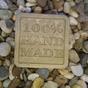 May include: A brown rectangular bar of soap with the text "100% HAND MADE" embossed on the front. The soap is sitting on a bed of small, smooth, gray and brown stones.