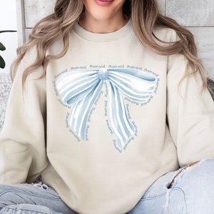 May include: A light beige sweatshirt with a blue watercolor bow design. The bow is made up of multiple ribbons with the word "Pharmacist" written on each ribbon.