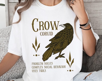 May include: White t-shirt featuring a detailed illustration of a crow in shades of brown and gold. The text "CROW CORVID" is arched above the bird, with additional text below stating "PROBLEM SOLVES COMPLEX SOCIAL BEHAVIOR USES TOOLS".