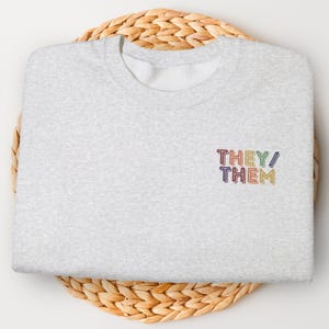 May include: A light gray sweatshirt with the embroidered text "THEY/ THEM" in rainbow colors. The sweatshirt is folded and placed on a round, woven, light brown placemat. The design is a statement of gender identity.