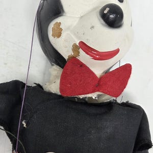 May include: A vintage Mickey Mouse puppet with black ears and head, a white face, and a red bow tie. The puppet has a black shirt and orange trousers with two white button details. The puppet shows signs of wear and tear.